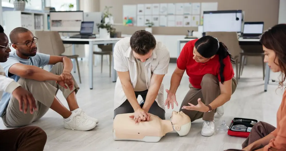 A group of office employees participating in a first aid training session, with an instructor demonstrating CPR techniques on a medical mannequin.
